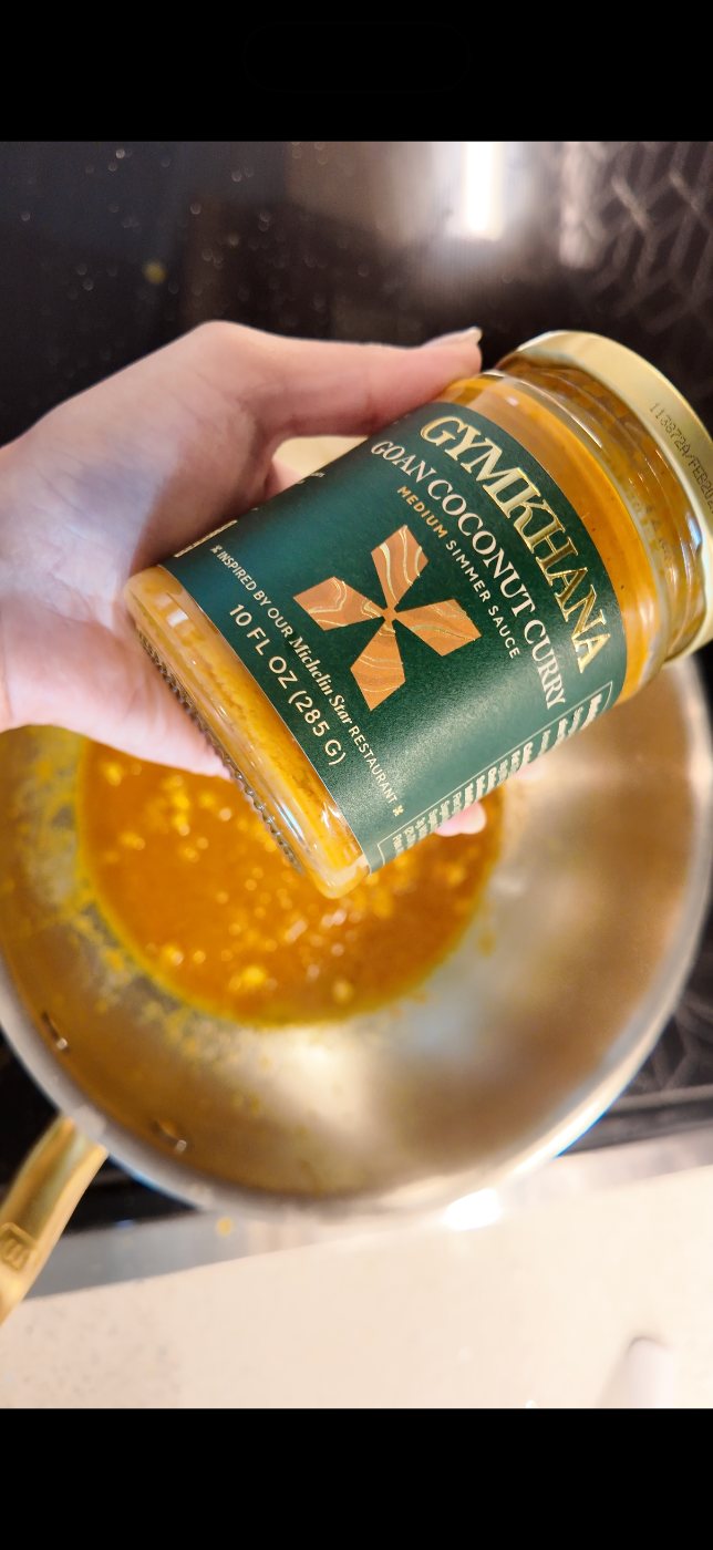 Jar of Gymkhana Goan Coconut Curry held over a pan of simmering sauce